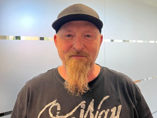 Mann mit Kinnbart, Cap und schwarzem Shirt vor einer matten Glaswand