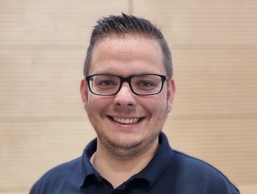 Grinsender Mann mit Brille, dunkelblonden, kurzen Haaren und einem blauen Poloshiert